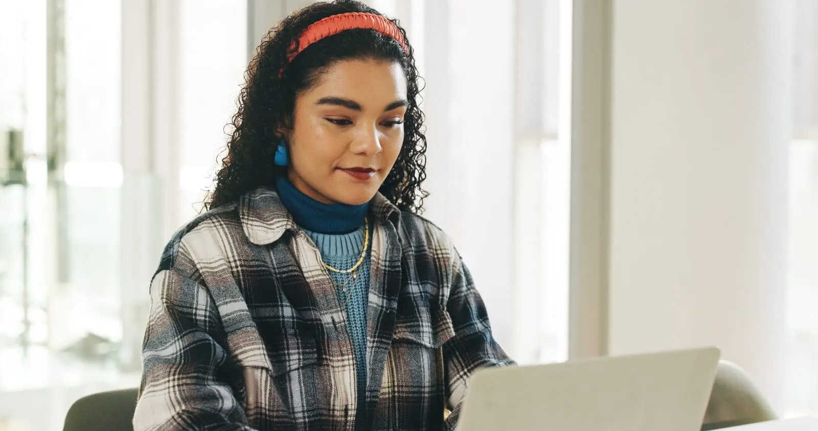 Woman in a plaid shirt working on a laptop, focused on reading and reviewing work at a desk.