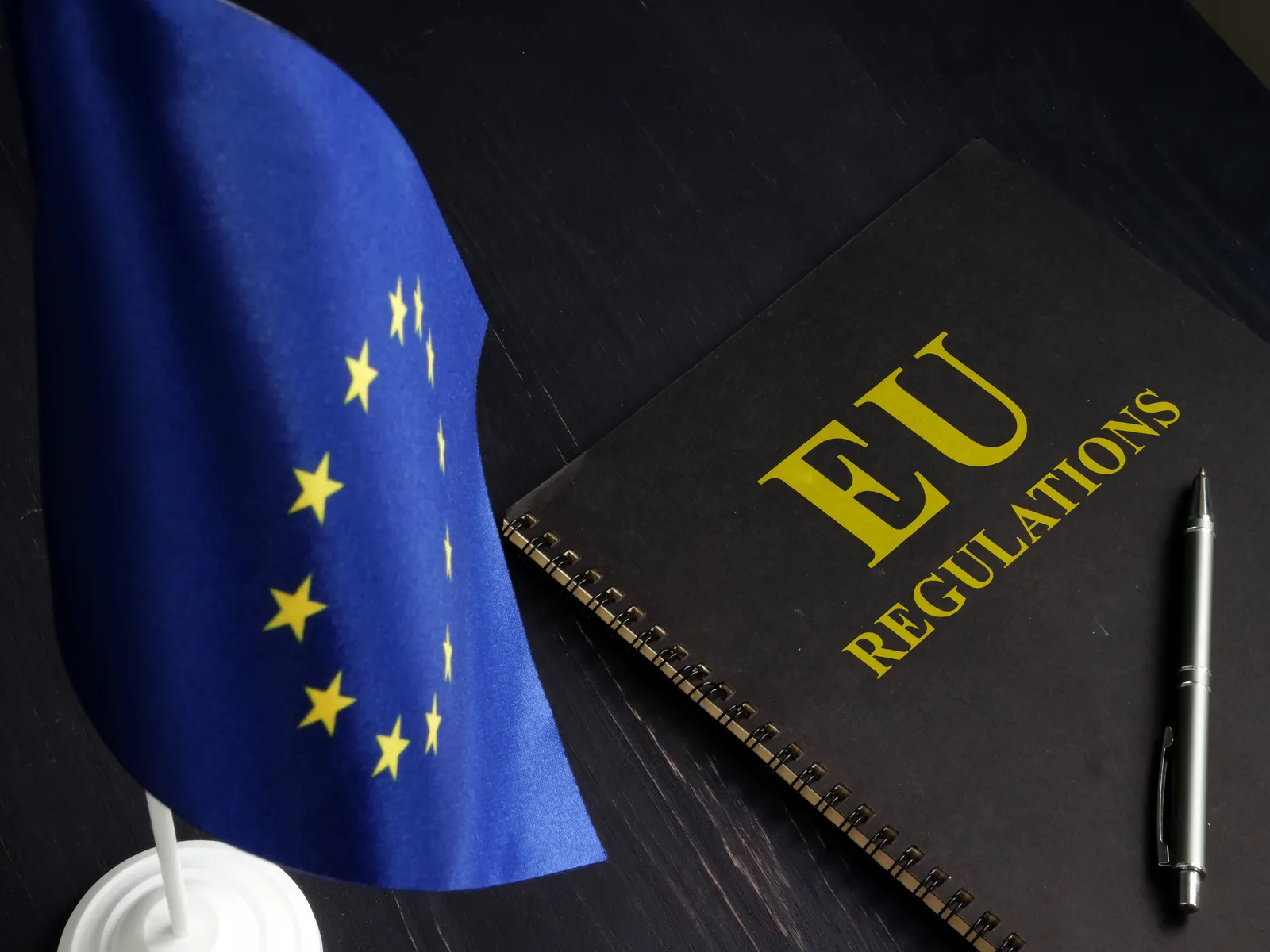 EU flag next to a black notebook labeled 'EU Regulations' with a pen on a dark desk, symbolizing European Union laws and policies.