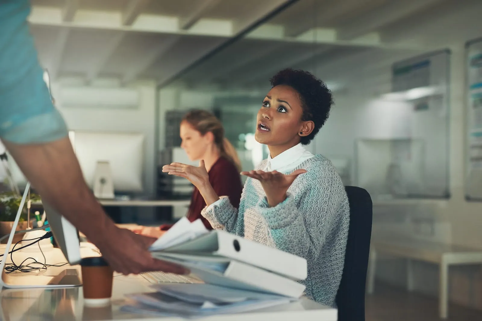 A frustrated woman sitting at her office desk raises her hands in exasperation while looking up. She appears overwhelmed as a colleague hands her a large stack of binders and paperwork.