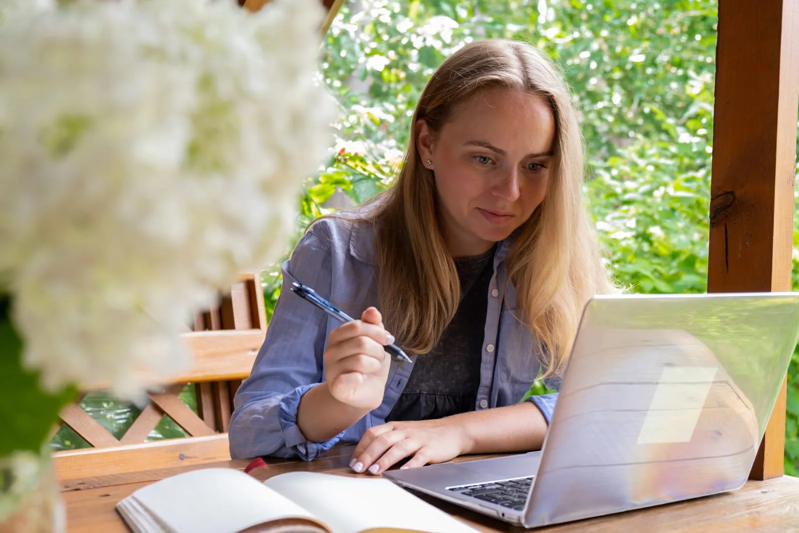 Young woman sitting at an outdoor wooden table, taking notes while working on a laptop in a natural setting with greenery.