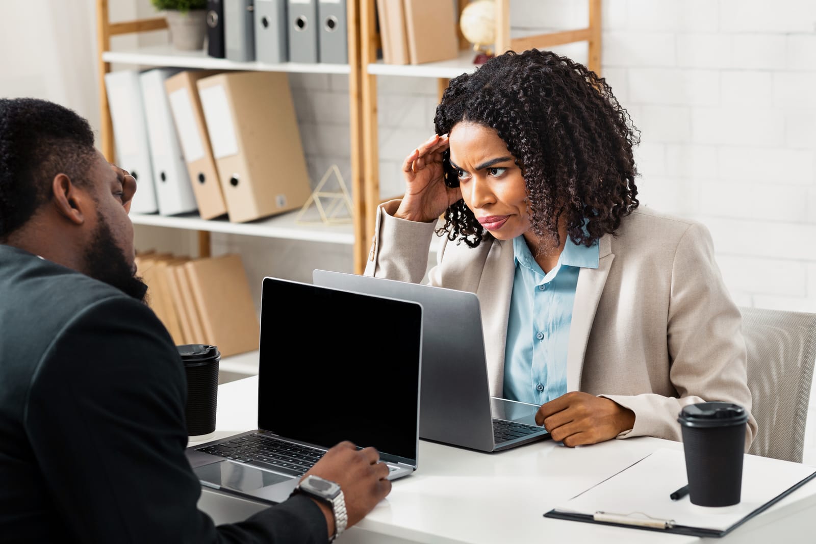 Two African American colleagues in an office, discussing work with laptops open; one woman appears frustrated while listening to her coworker.