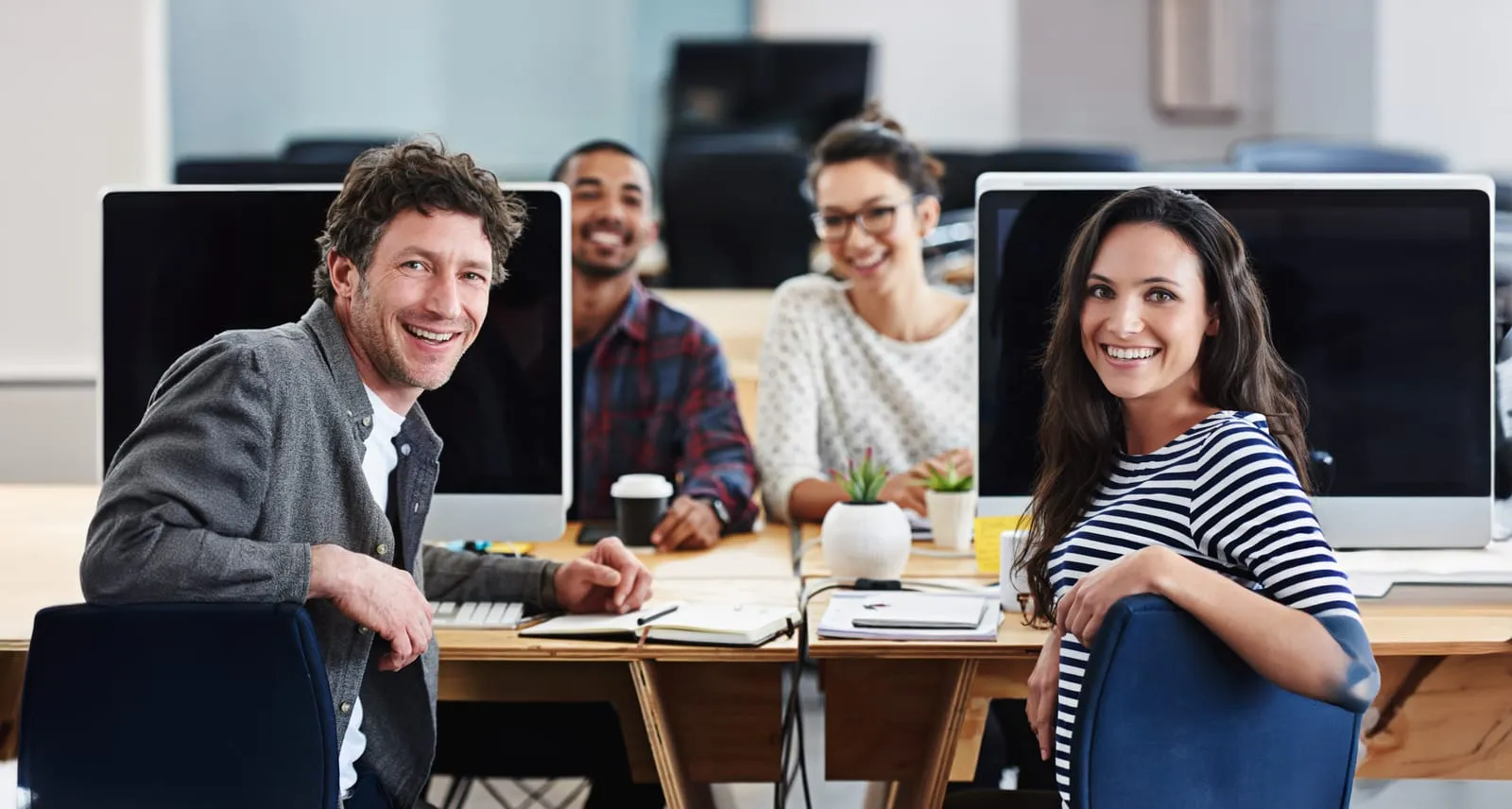 A group of four young professionals is sitting at a modern, open-concept office workspace, smiling and looking at the camera. Two individuals in the foreground, a man in a gray shirt and a woman in a striped shirt, are leaning slightly towards each other, appearing engaged and happy. Behind them, two colleagues, a man in a plaid shirt and a woman in glasses, are also smiling.
