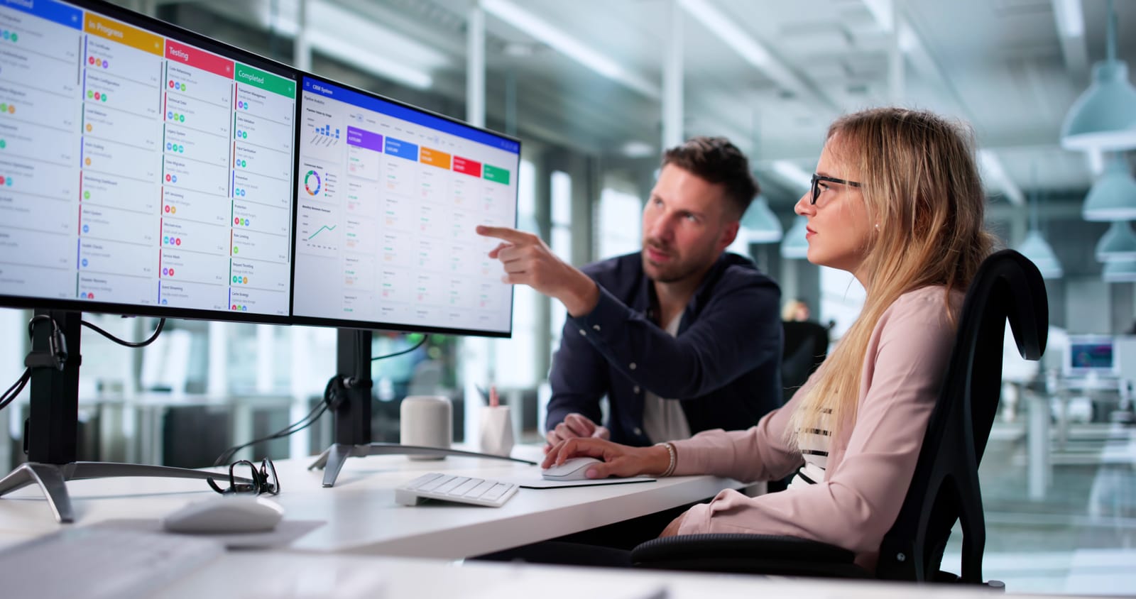 Man and woman working together at a desk in a modern office, analyzing data on large computer screens with project management software.