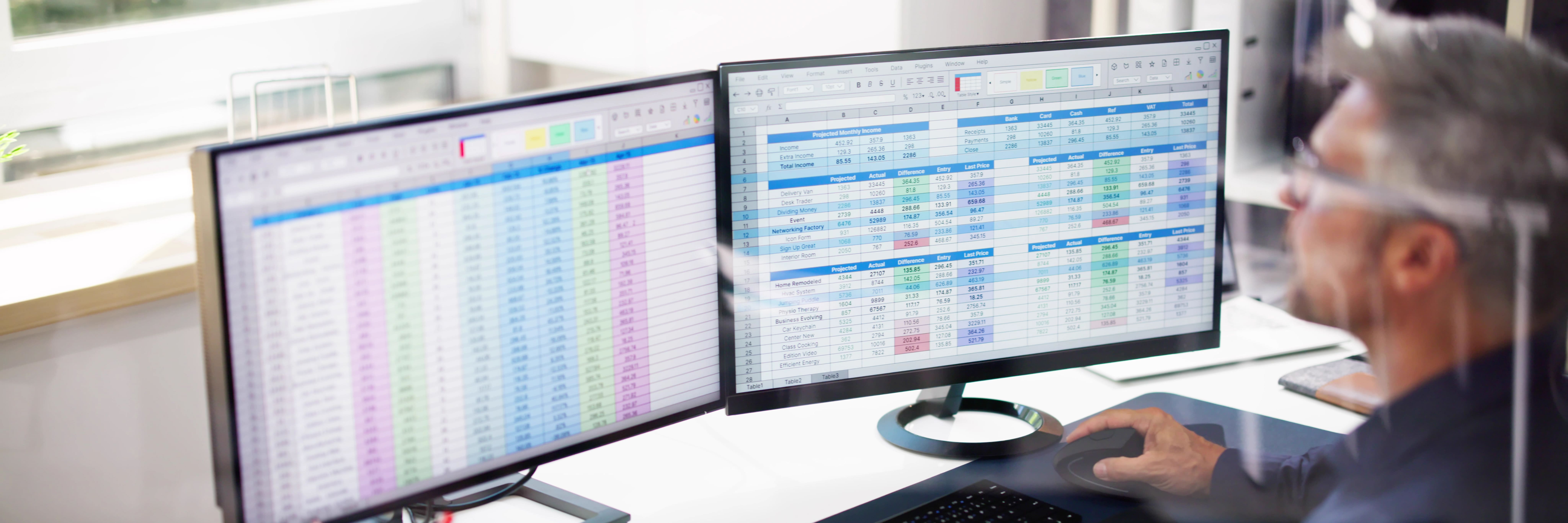 Man working on dual monitors reviewing large spreadsheets with financial data in an office setting