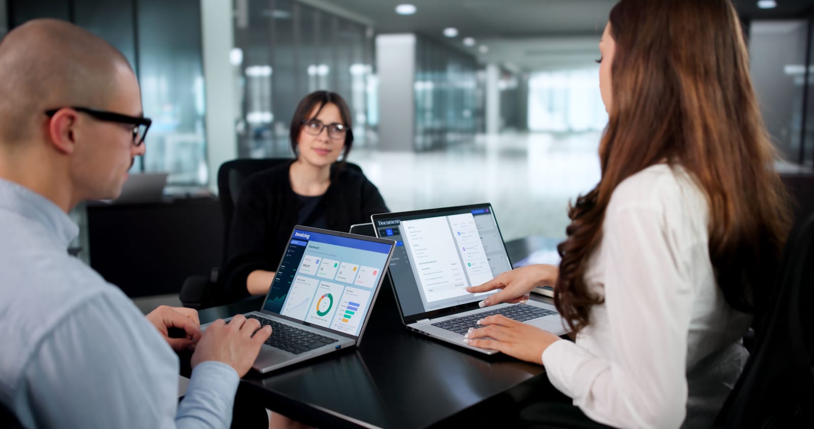 Three professionals collaborating in a modern office, reviewing data dashboards on laptops during a meeting.