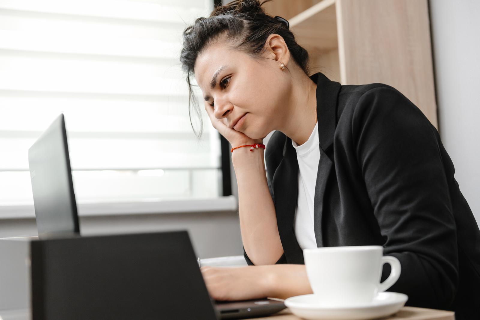 An office worker sitting at a desk, looking at a laptop screen with a cup of coffee nearby