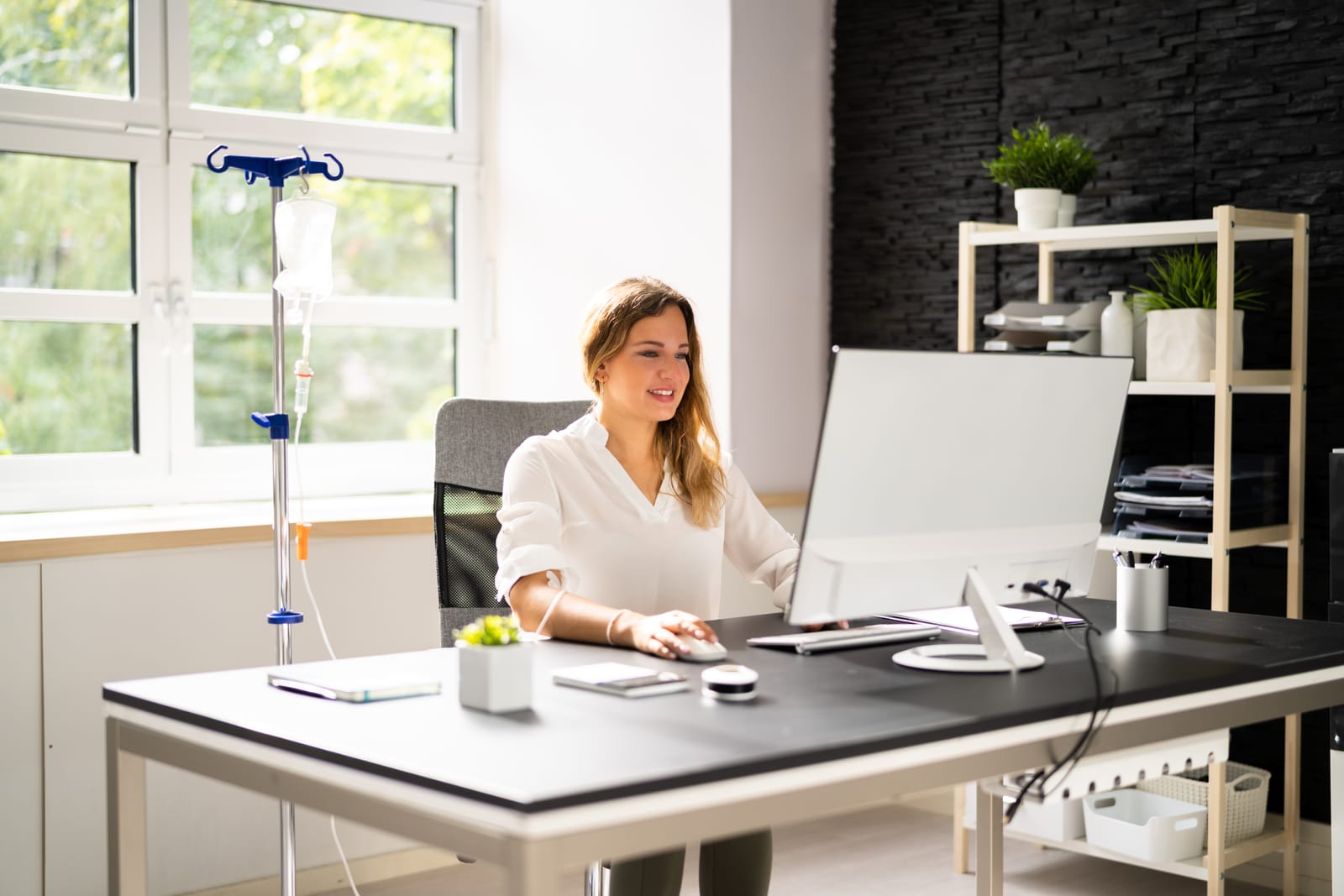 Woman receiving IV drip therapy while working at a computer in a bright modern office.