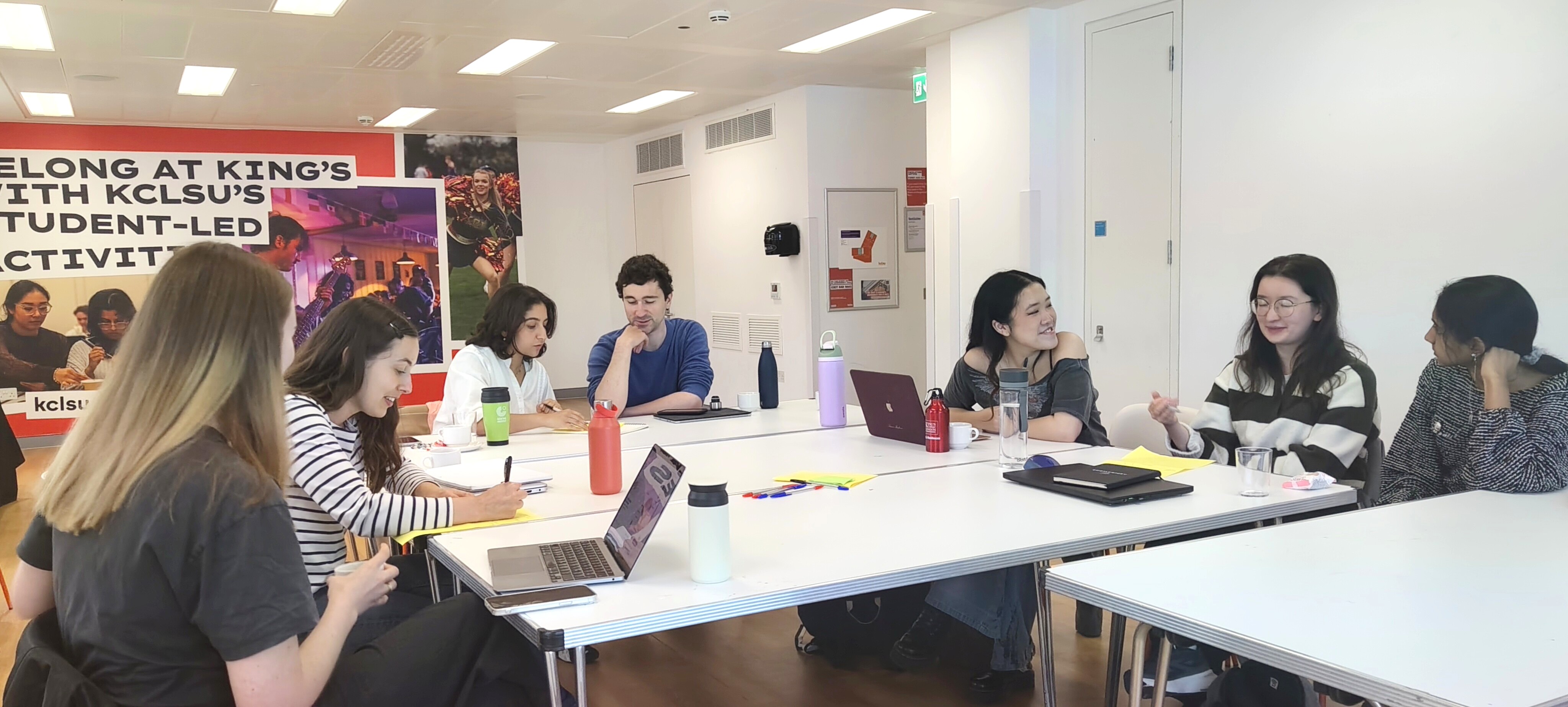 Seven students sitting around a large white table with laptops and books. They are smiling and talking. They're sitting in a bright white room and some are drawing on green and yellow paper.