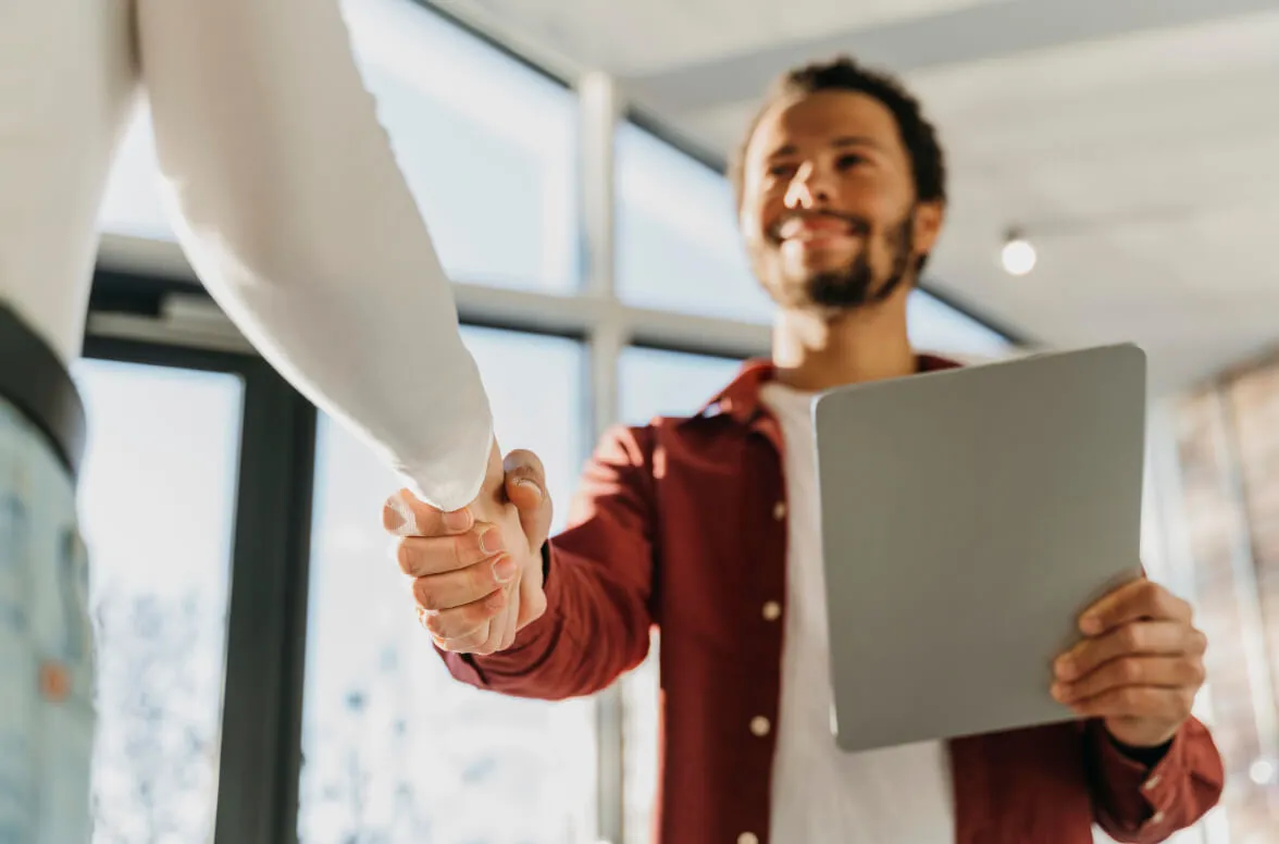 Dos personas estrechando las manos en una oficina, uno sostiene una tableta y sonríe.