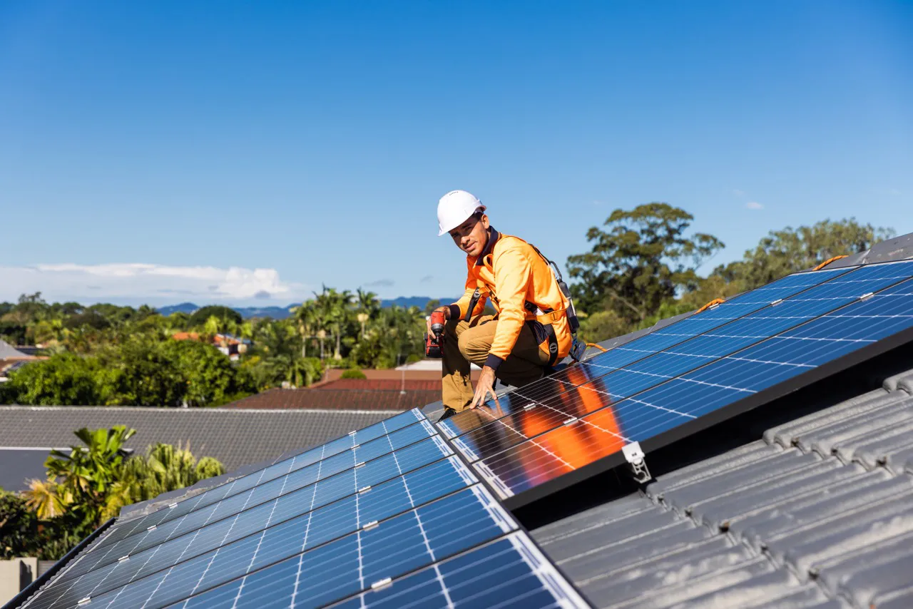 Trabajador con casco blanco y chaleco naranja instalando paneles solares en un techo bajo un cielo despejado.