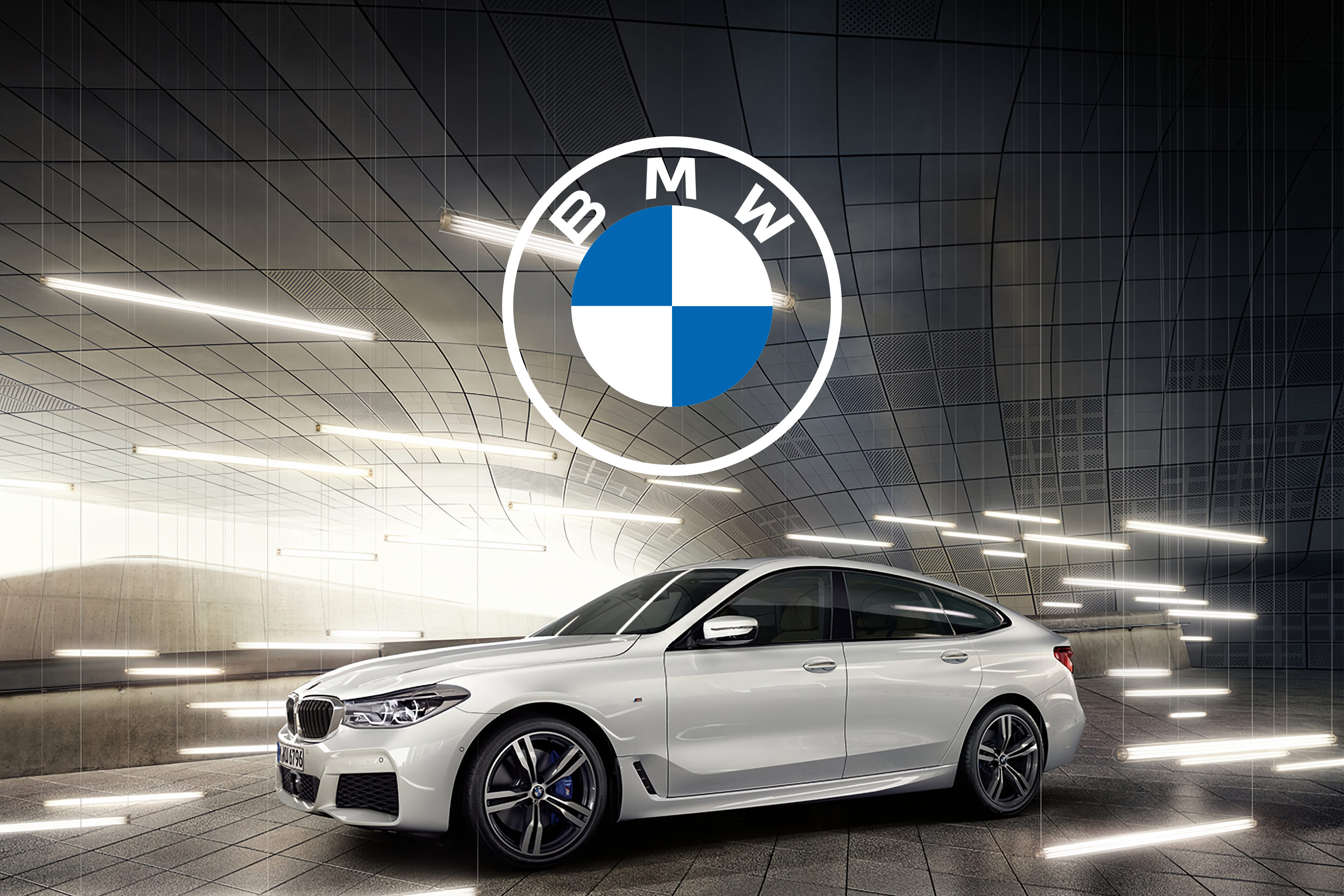 BMW : le logo de la marque au-dessus d’une BMW blanche, dans un tunnel voûté traversé de tubes de lumière