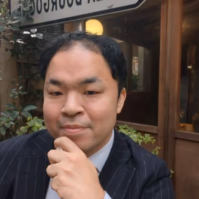 Man in a dark pinstripe suit with a light blue shirt resting his chin on his hand, sitting outdoors near a wooden window and plants.