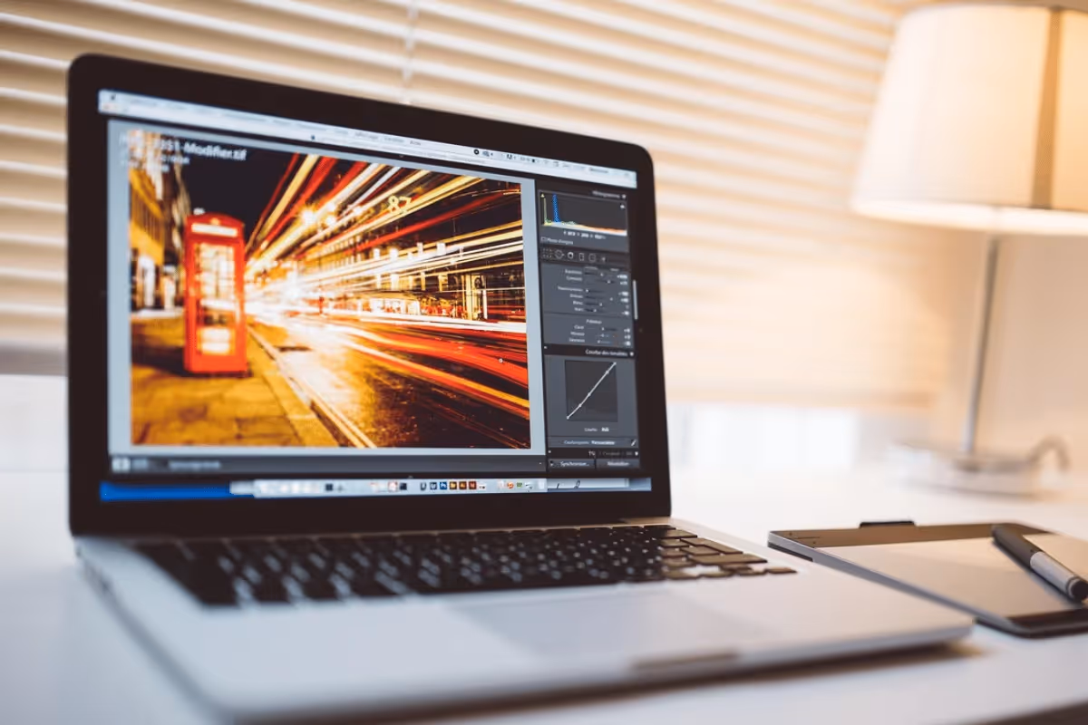 A laptop displaying a vibrant photo with light trails, next to a tablet and pen, set in a well-lit workspace with blinds.