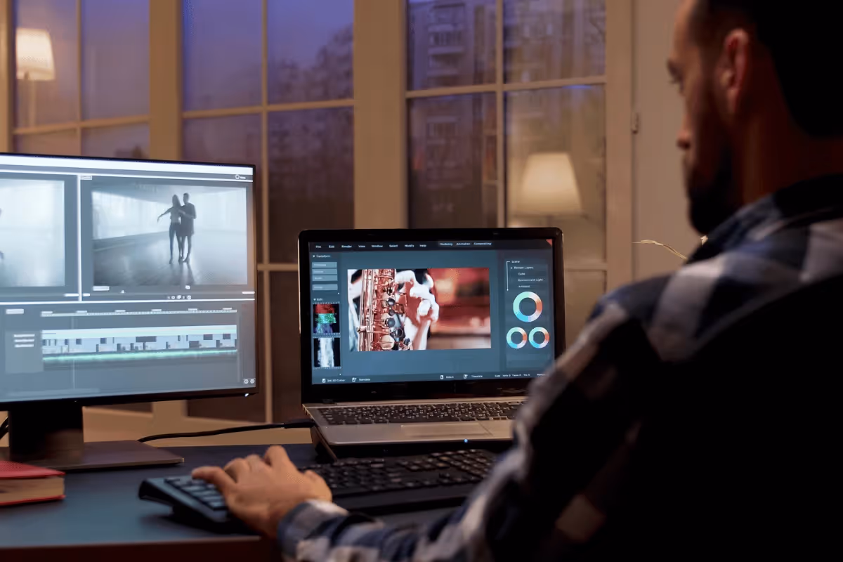 A person edits video on two screens at a dimly lit desk, with a nighttime city view through a window behind them.