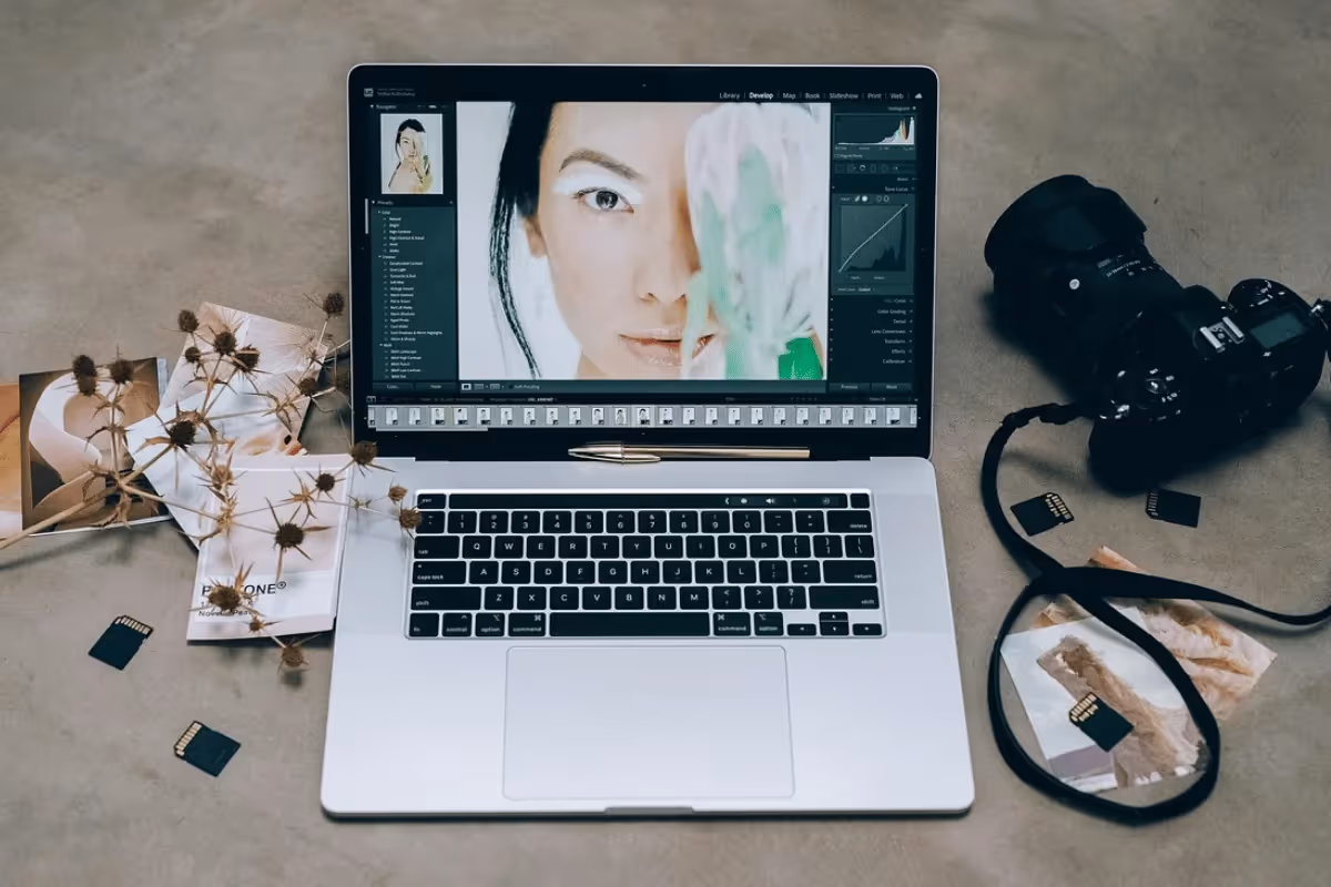 A laptop with photo editing software open, surrounded by SD cards, printed photos, a camera, and dried flowers on a neutral background.
