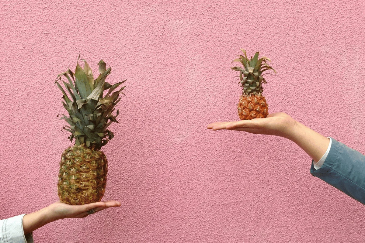 Two hands hold pineapples against a pink textured background. The left hand holds a large pineapple, the right hand a smaller one, creating a playful contrast.