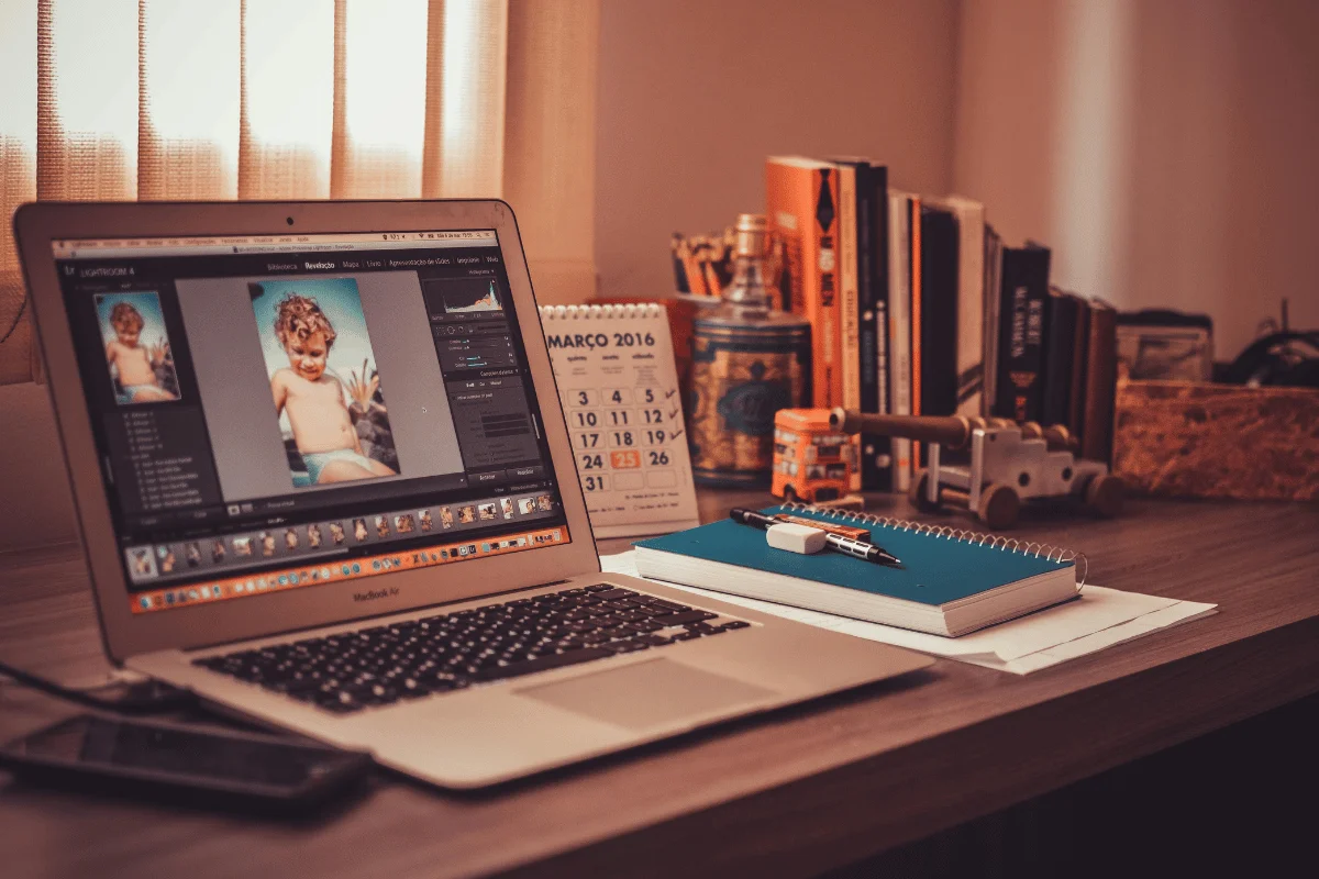 A laptop on a wooden desk displays photo editing software, surrounded by books, a calendar, a notebook, and stationery items.