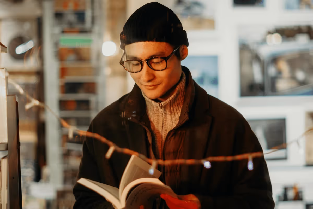 A person wearing glasses, a beanie, and a coat reads a book in a warmly lit room. The background features blurred photos, adding a cozy ambiance.