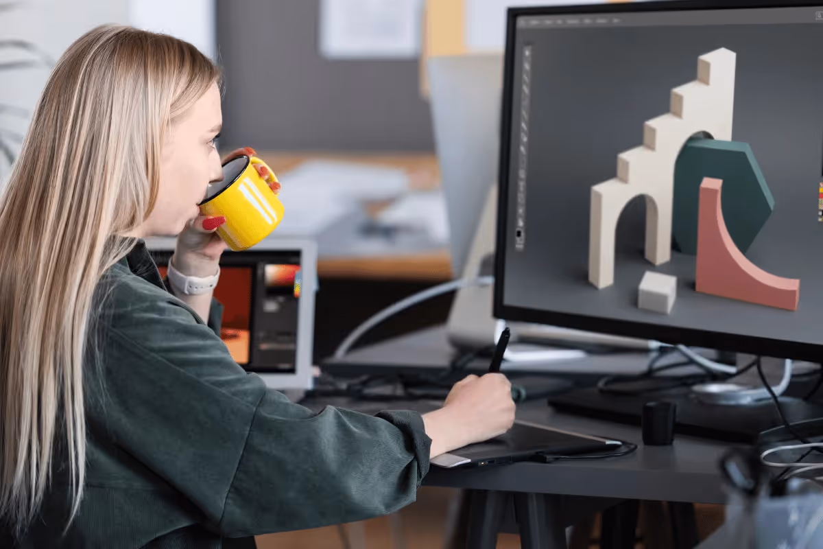 A freelancer works at a desk using a graphics tablet and a computer displaying 3D design models, holding a yellow mug in one hand.
