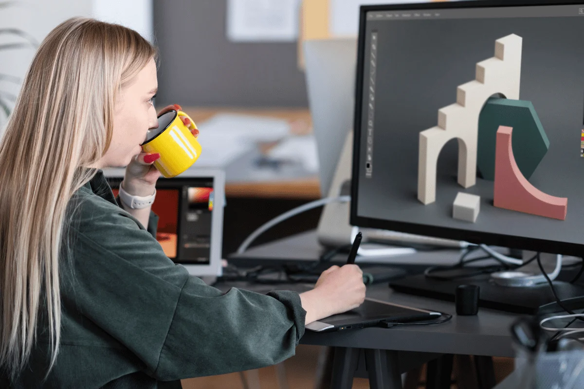 A freelancer works at a desk using a graphics tablet and a computer displaying 3D design models, holding a yellow mug in one hand.