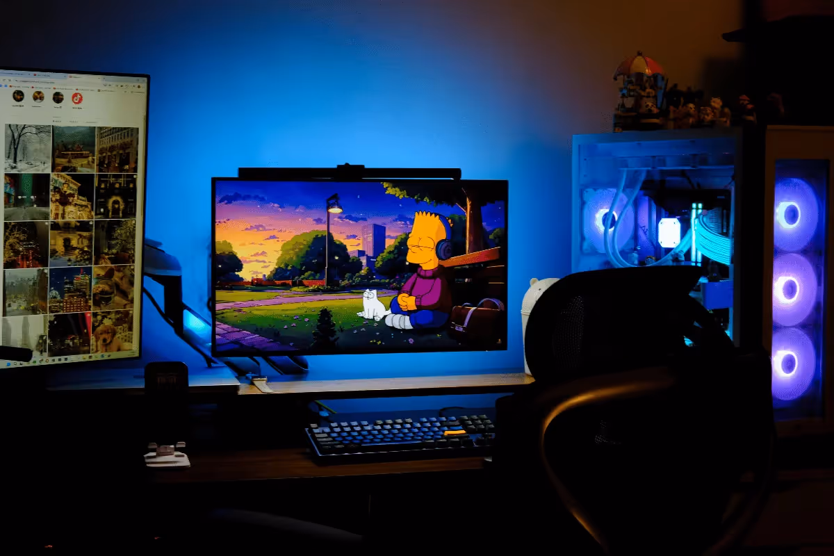 A modern workspace featuring two monitors; one displays Bart Simpson in a colorful sunset scene, with RGB lighting and a gaming chair present.