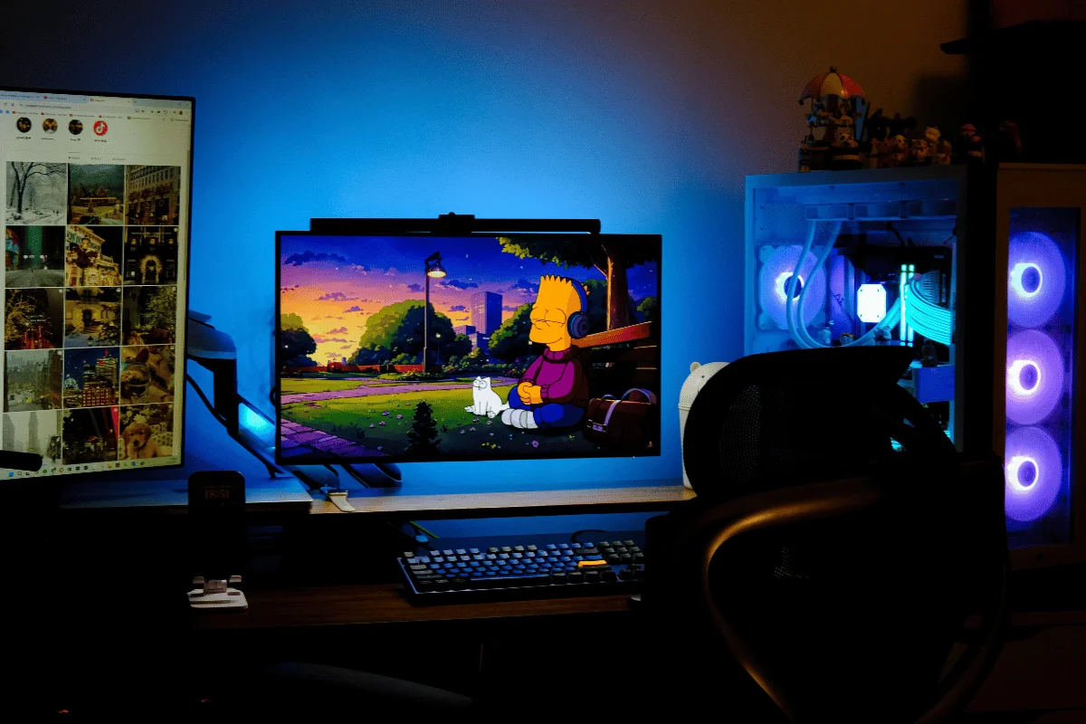 A modern workspace featuring two monitors; one displays Bart Simpson in a colorful sunset scene, with RGB lighting and a gaming chair present.