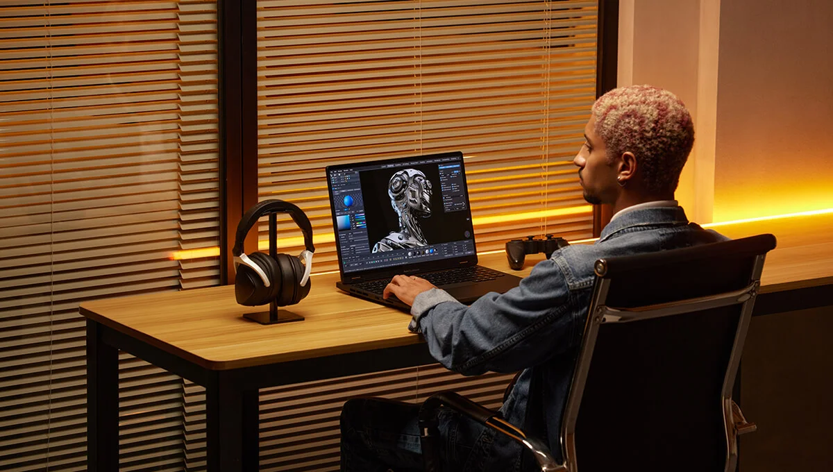 A person sits at a sleek desk, working on a laptop displaying a detailed robot model, with headphones and a game controller nearby.
