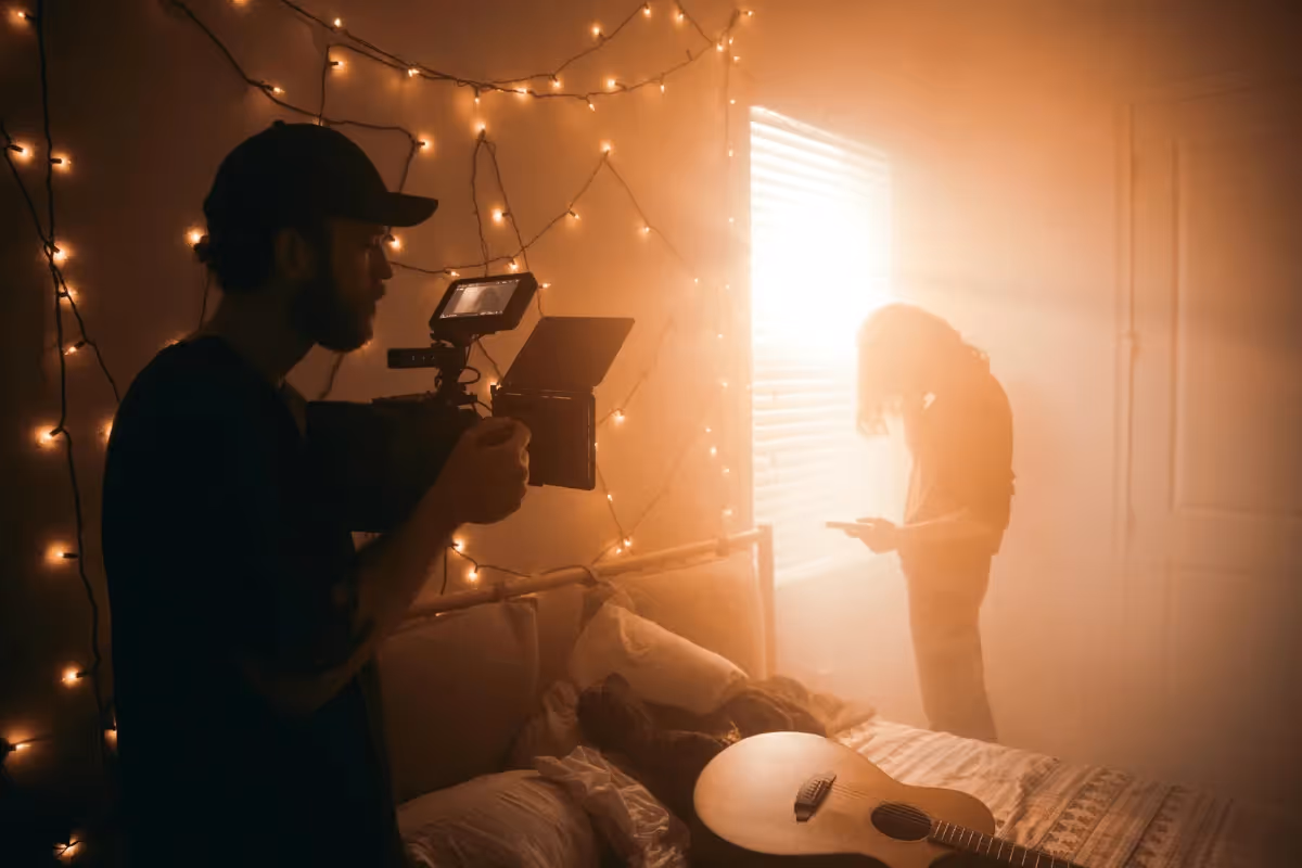 A filmmaker with a camera captures a person near a sunny window, in a cozy room illuminated by string lights. A guitar rests on the bed, creating a calm, artistic atmosphere.
