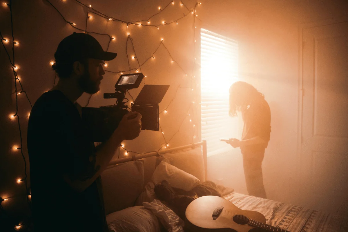 A filmmaker with a camera captures a person near a sunny window, in a cozy room illuminated by string lights. A guitar rests on the bed, creating a calm, artistic atmosphere.