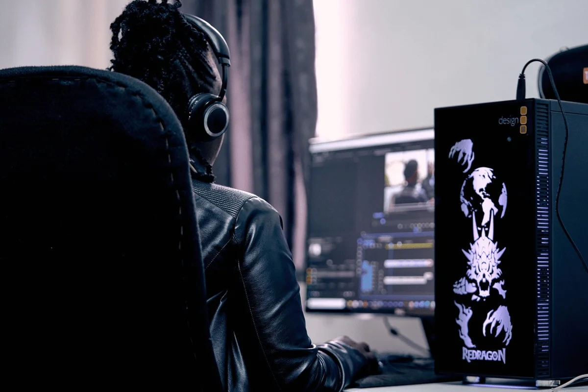 A person in a leather jacket and headphones edits a video on dual monitors. A computer with a fantasy-themed design is visible, conveying focus and creativity.