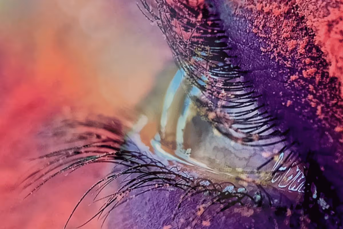Close-up of an eye with colorful eyeshadow and mascara, surrounded by vibrant gradients of purple and orange. The mood is artistic and dramatic.