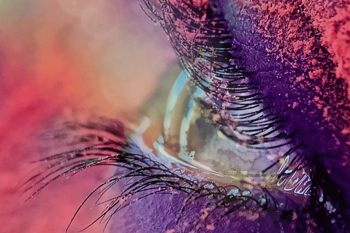 Close-up of an eye with colorful eyeshadow and mascara, surrounded by vibrant gradients of purple and orange. The mood is artistic and dramatic.