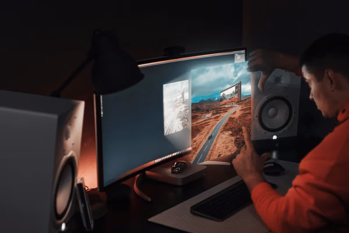 A man in a red sweater uses a computer with a large curved monitor displaying a digital landscape. The room is dimly lit, conveying a focused, creative ambiance.