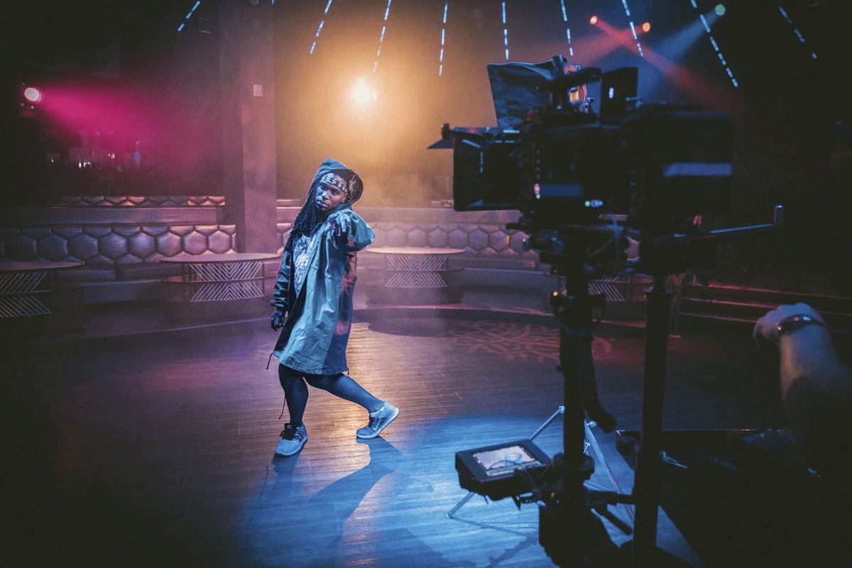 A dancer strikes a pose on stage, illuminated by colorful lights, with a camera capturing the dynamic performance in a nightclub setting.
