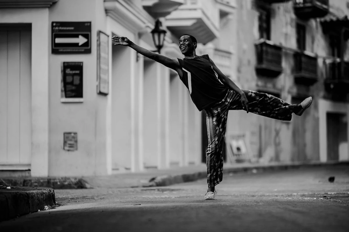 A joyful dancer is mid-motion on an empty street, extending one leg and arm, exuding happiness and energy. The scene is in black and white.