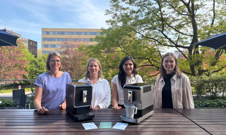 Four women stand behind two Tchibo coffee machines, presented as part of a take-back pilot project in collaboration with koorvi. Informational cards on sustainable consumption are placed on the table. In the background: spring greenery on a sunny terrace.