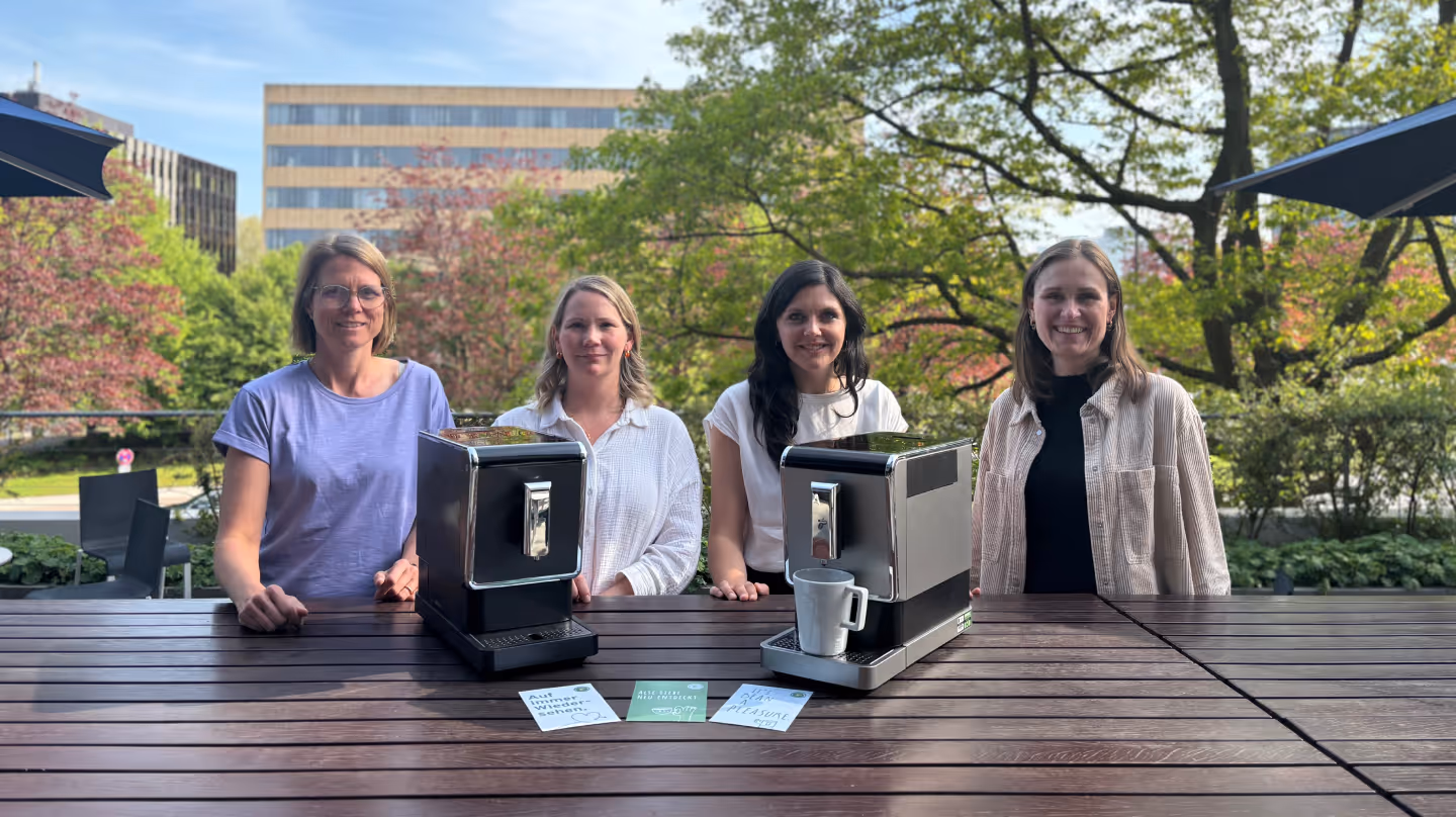 Four women stand behind two Tchibo coffee machines, presented as part of a take-back pilot project in collaboration with koorvi. Informational cards on sustainable consumption are placed on the table. In the background: spring greenery on a sunny terrace.