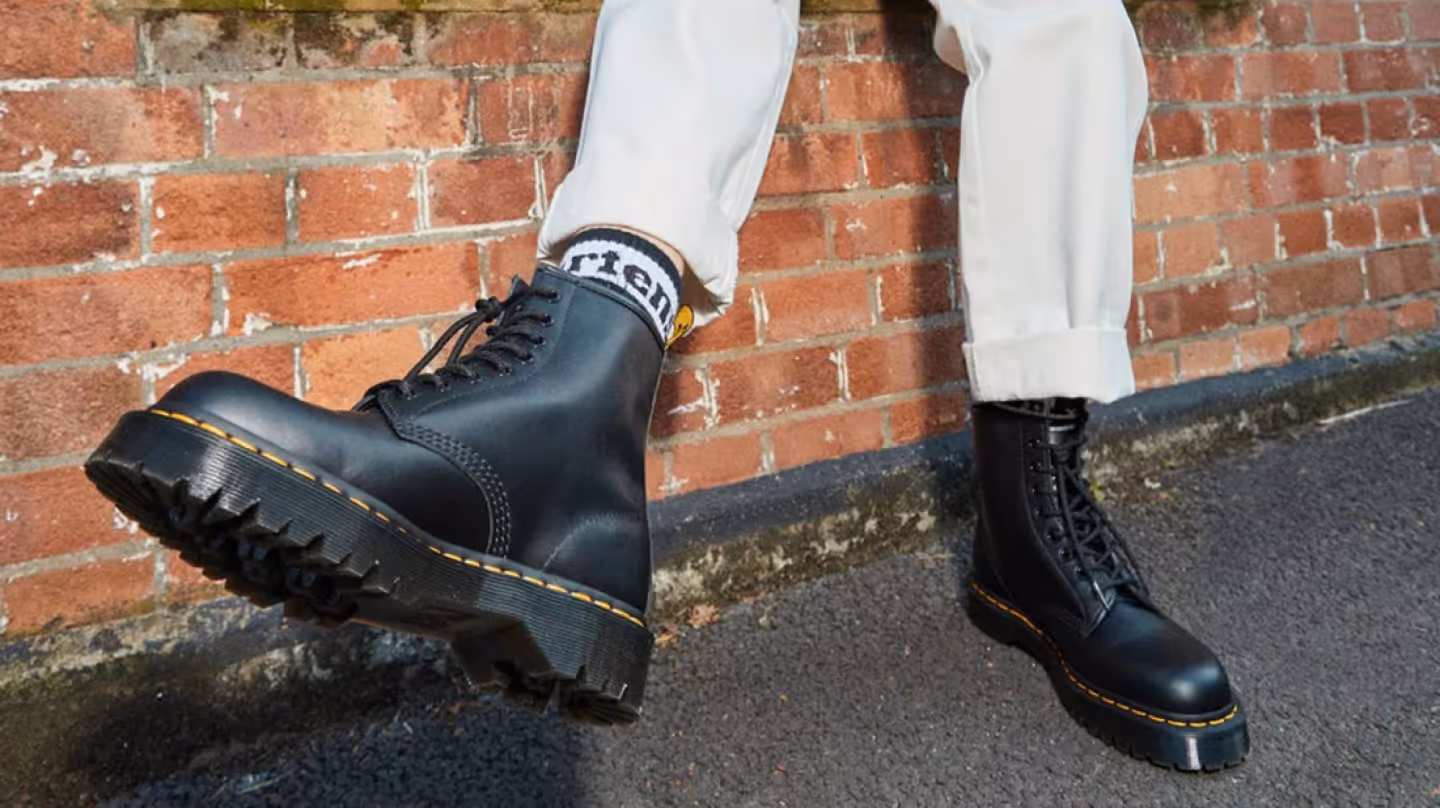 Schwarze Dr. Martens Boots getragen von einer Person vor einer Backsteinmauer.