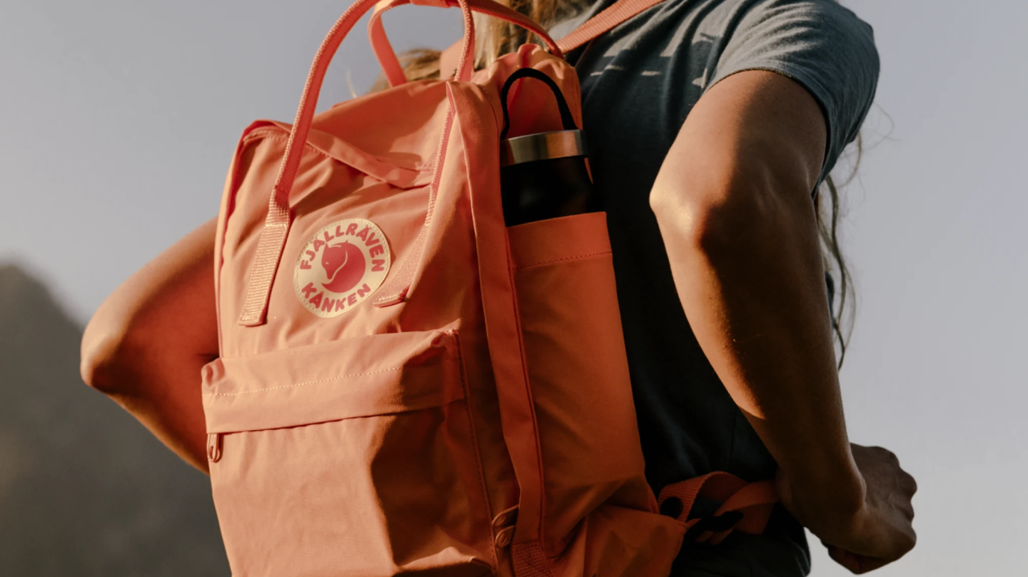 Nahaufnahme eines orangenen Fjällräven Kånken-Rucksacks mit Trinkflasche in der Seitentasche, getragen von einer Person im Freien.