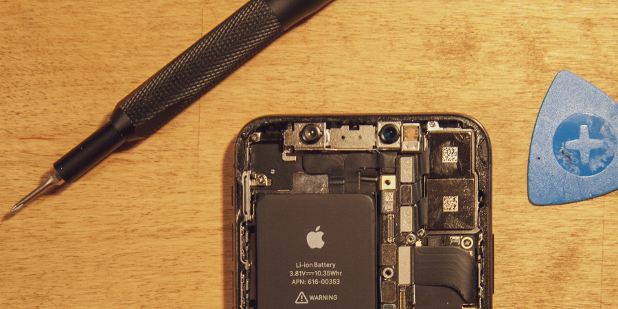 Close-up of a partially disassembled smartphone with visible battery and internal components, lying on a wooden surface next to a precision screwdriver and a blue plastic opening tool.