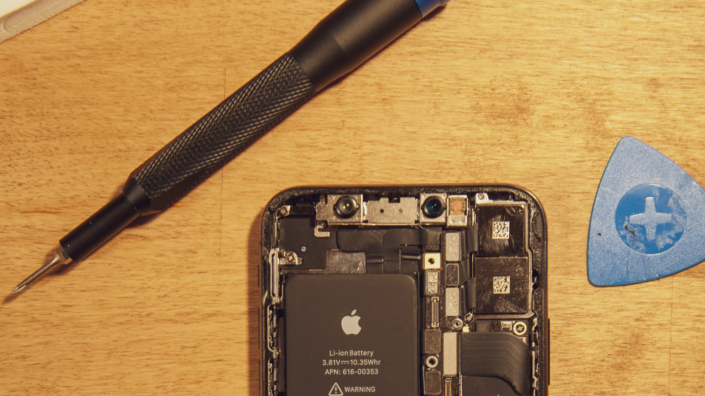 Close-up of a partially disassembled smartphone with visible battery and internal components, lying on a wooden surface next to a precision screwdriver and a blue plastic opening tool.