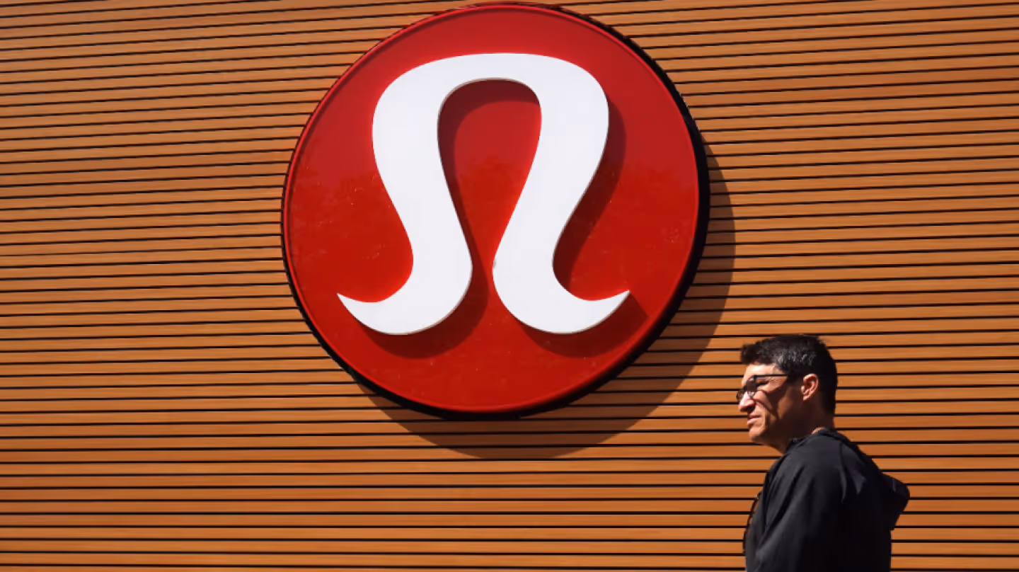 Exterior view of a Lululemon store with a large red and white logo on a wooden facade, and a man walking past.