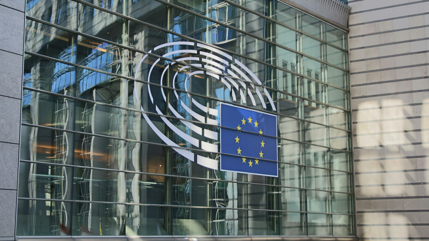 Glass facade of the European Parliament with EU flag and logo.