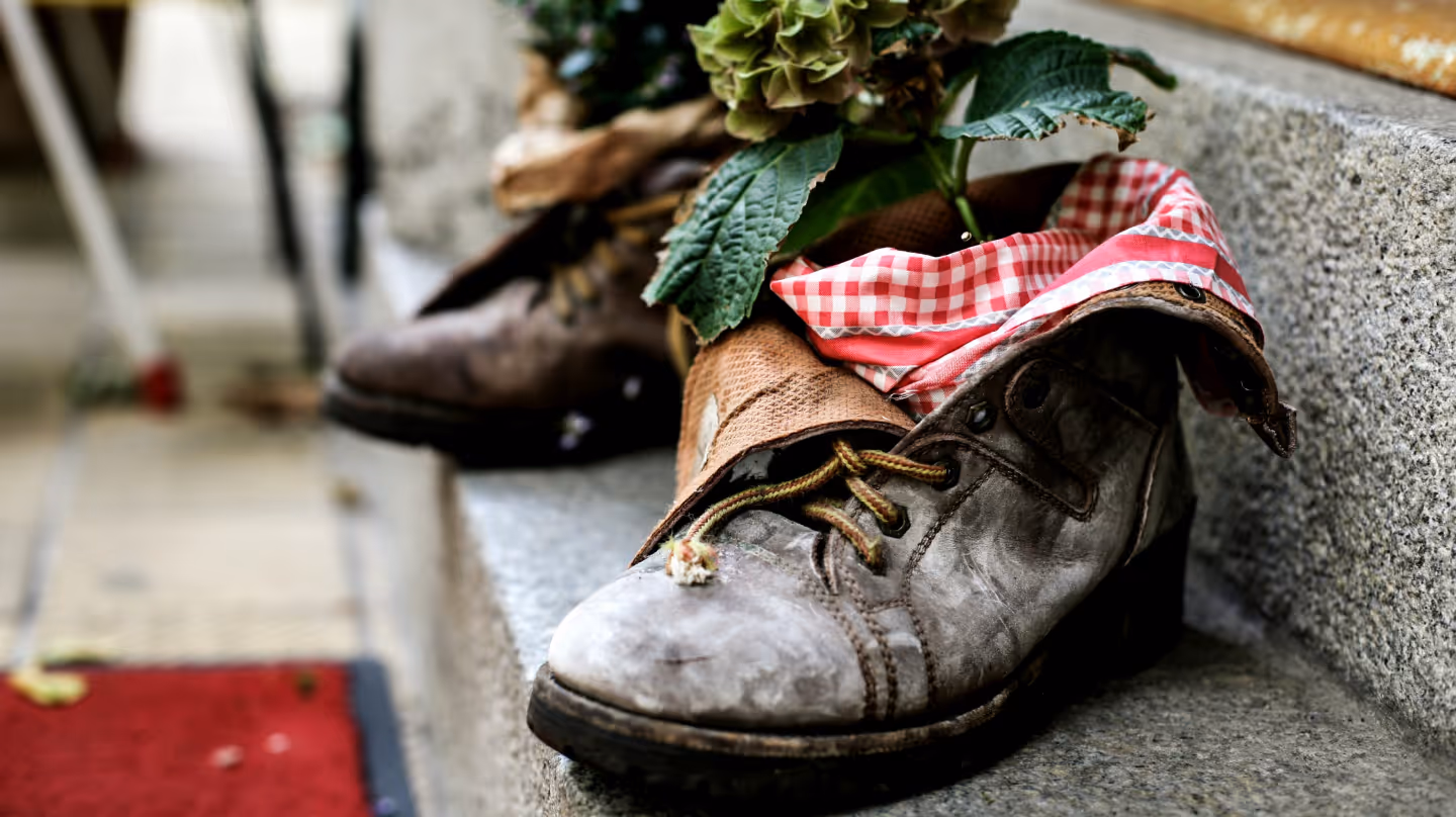 Old, worn-out boots creatively repurposed as flower pots with plants and red-and-white checkered fabric.