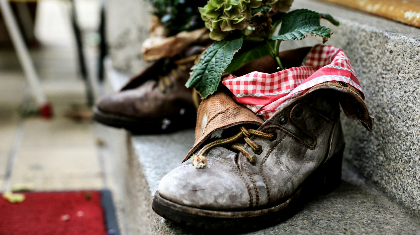 Old, worn-out boots creatively repurposed as flower pots with plants and red-and-white checkered fabric.