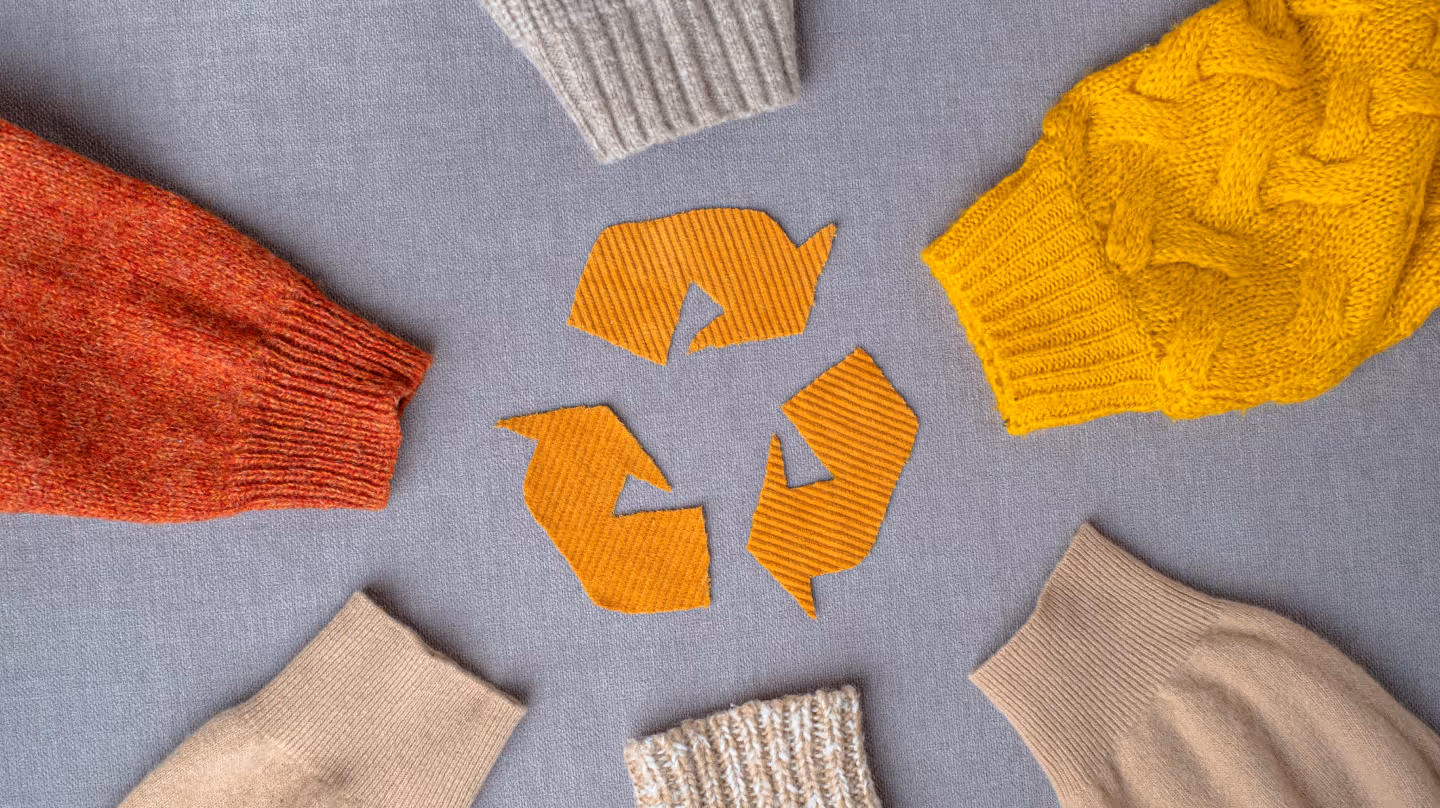 Colorful knitted sweaters arranged around orange recycling symbol on gray background representing textile circular economy