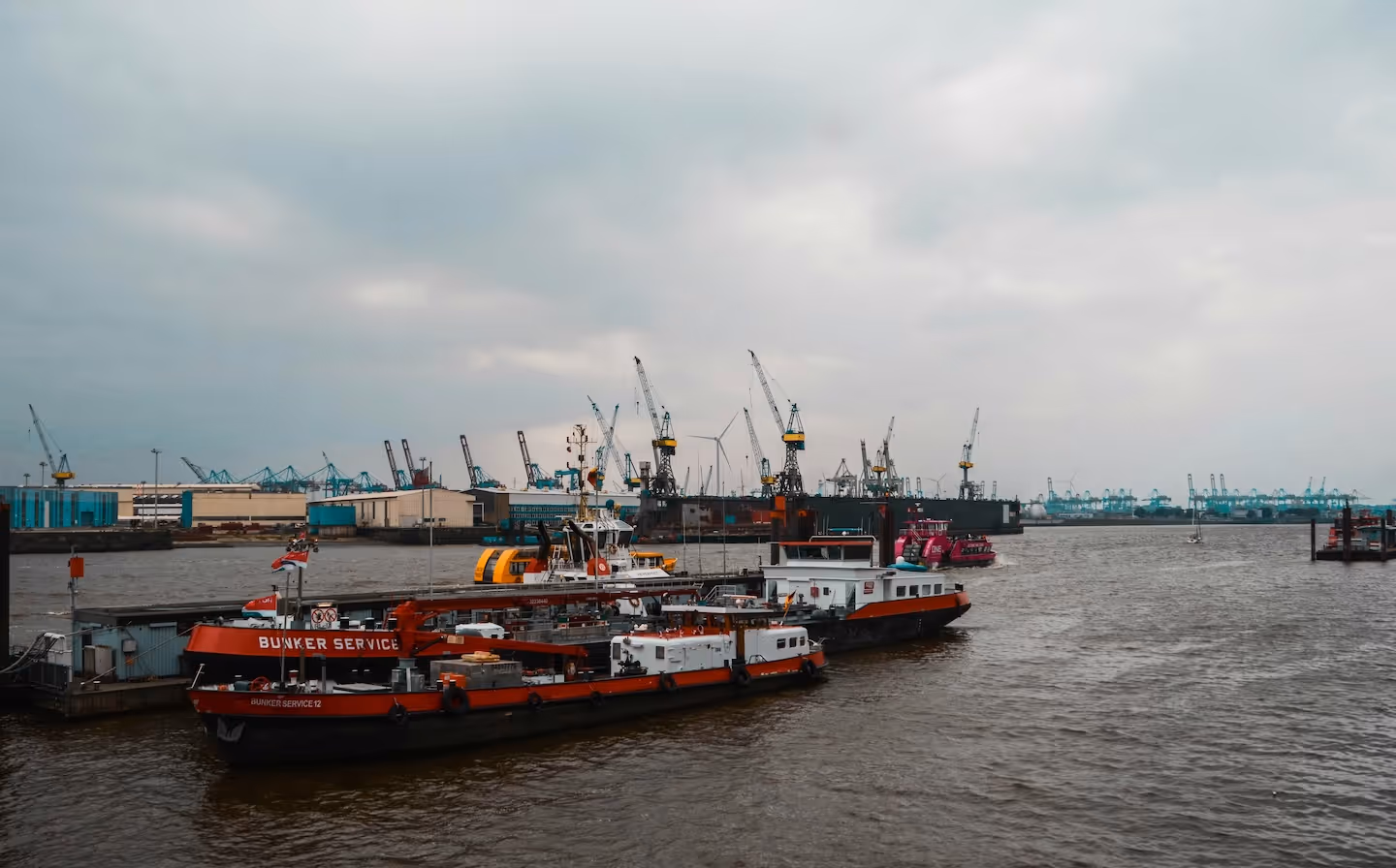 Red bunker service boats moored at Hamburg port with cargo cranes in the background under overcast sky