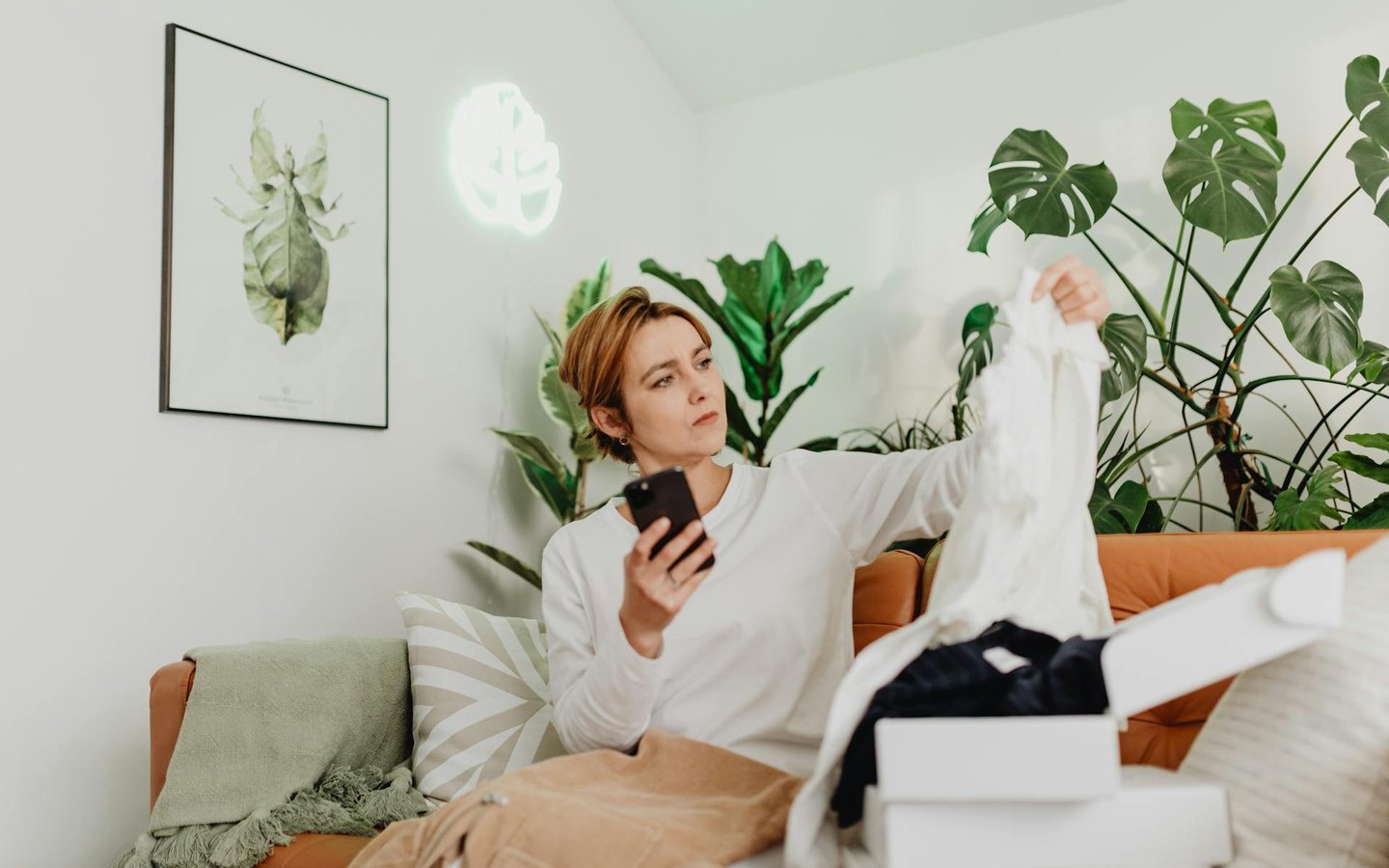 Woman on sofa inspecting clothing item while scrolling on her smartphone, representing secondhand shopping and the resale boom