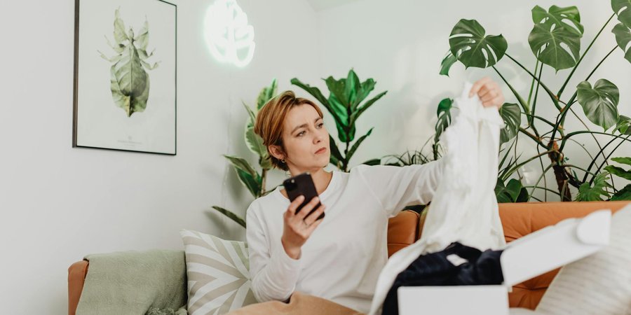 Woman on sofa inspecting clothing item while scrolling on her smartphone, representing secondhand shopping and the resale boom