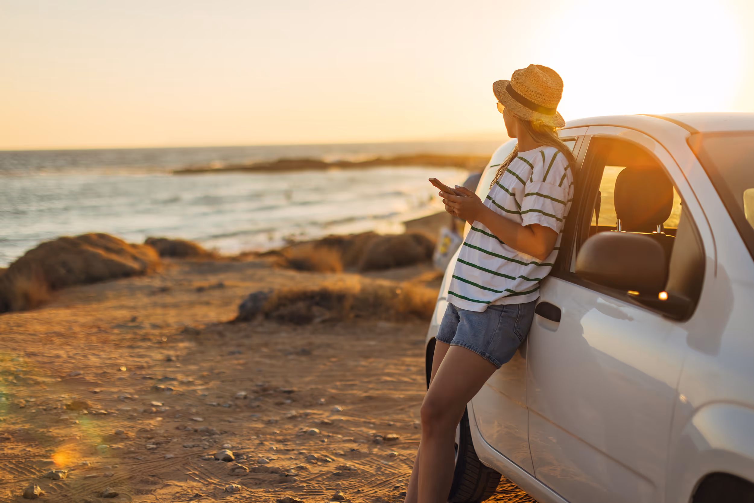 Woman in a striped shirt and hat leaning on a white car by the ocean at sunset holding a phone.